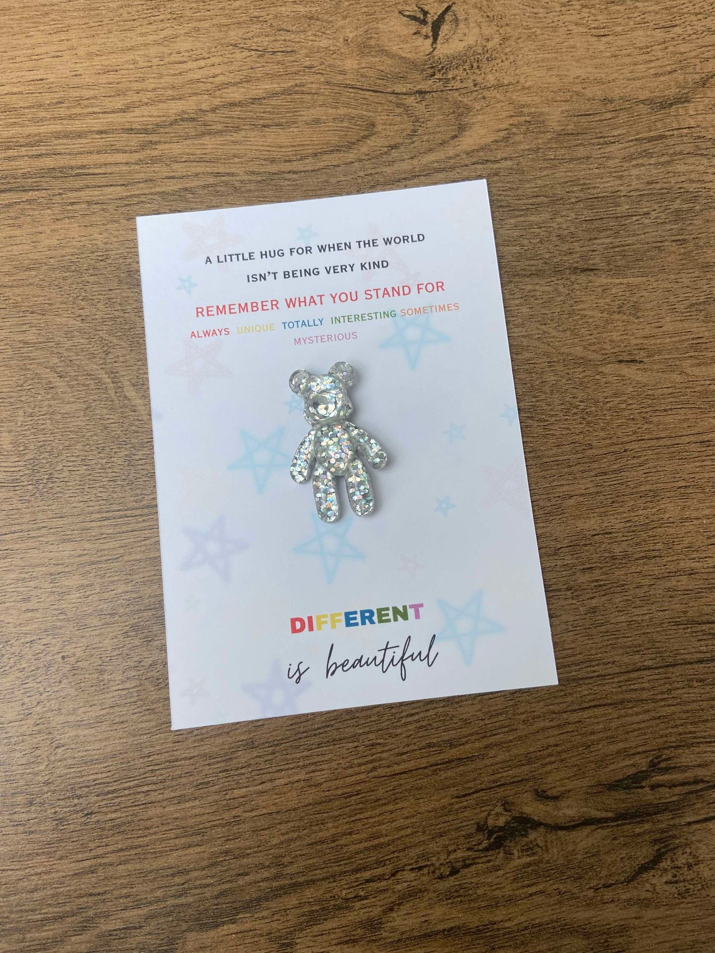 A multicolored teddy bear pocket hug with a note card that reads 'Autism Different Is Beautiful' and 'Remember what you stand for, not what you look like.' The bear is wearng a sparkly halo and the card is presented on a wooden surface.
