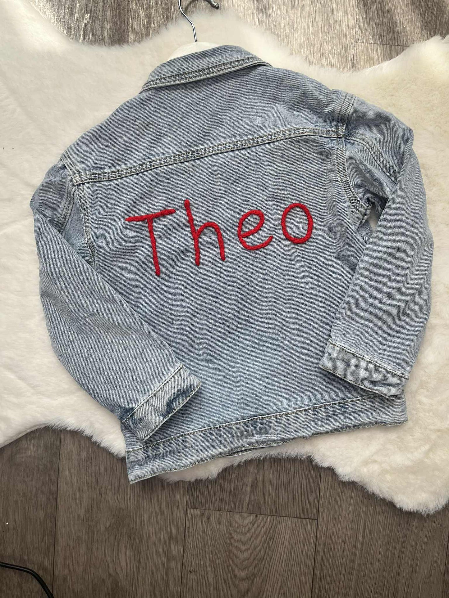 Denim jacket with 'Theo' embroidered on it, placed on a textured surface.