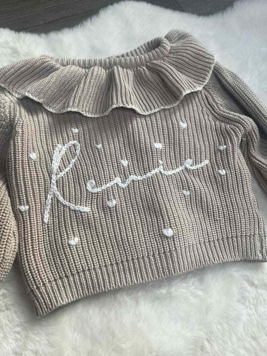 Gray ribbed sweater with ruffled collar and embroidered name on a fluffy white surface
