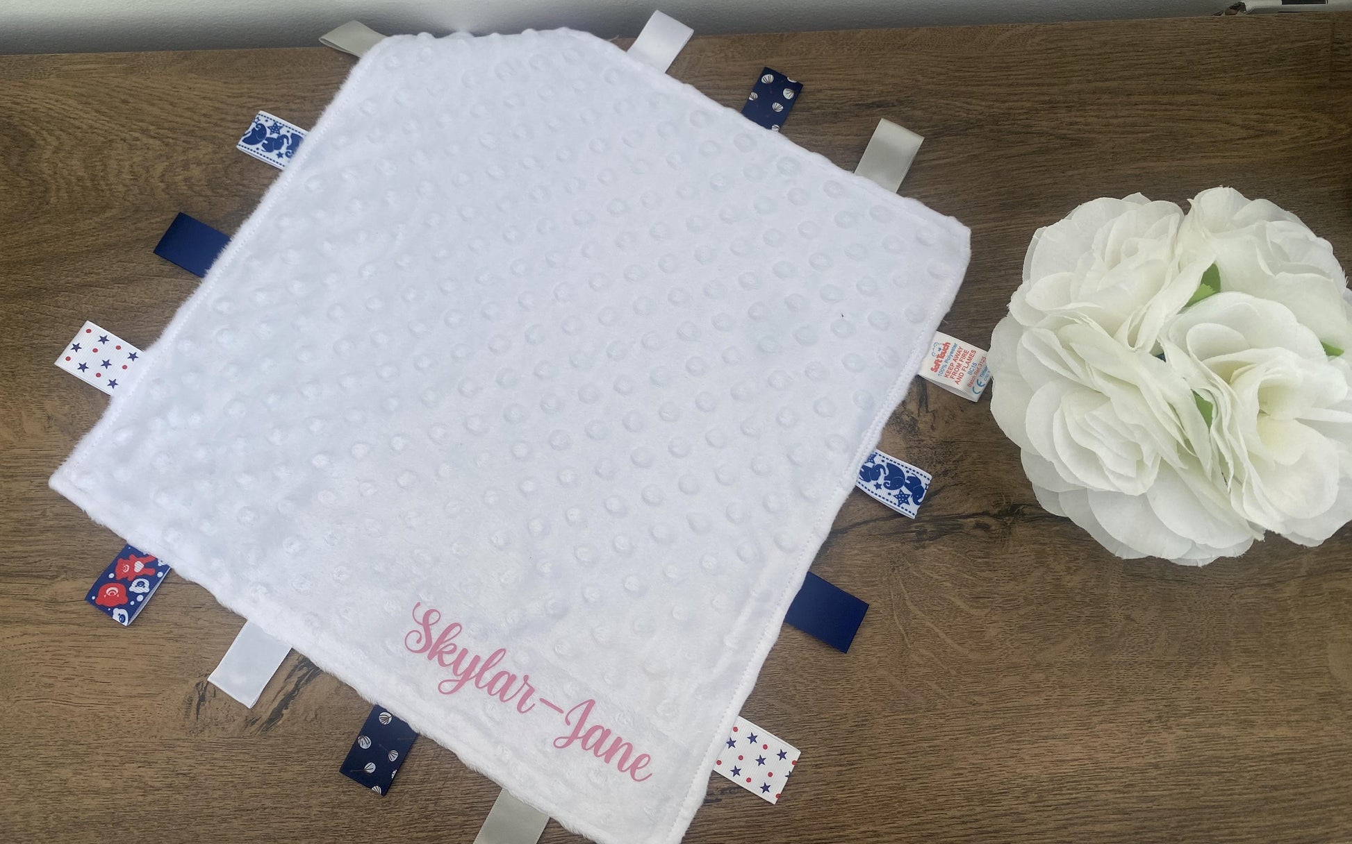 A personalised white baby comforter with blue and red patches, and a custom name printed in pink.