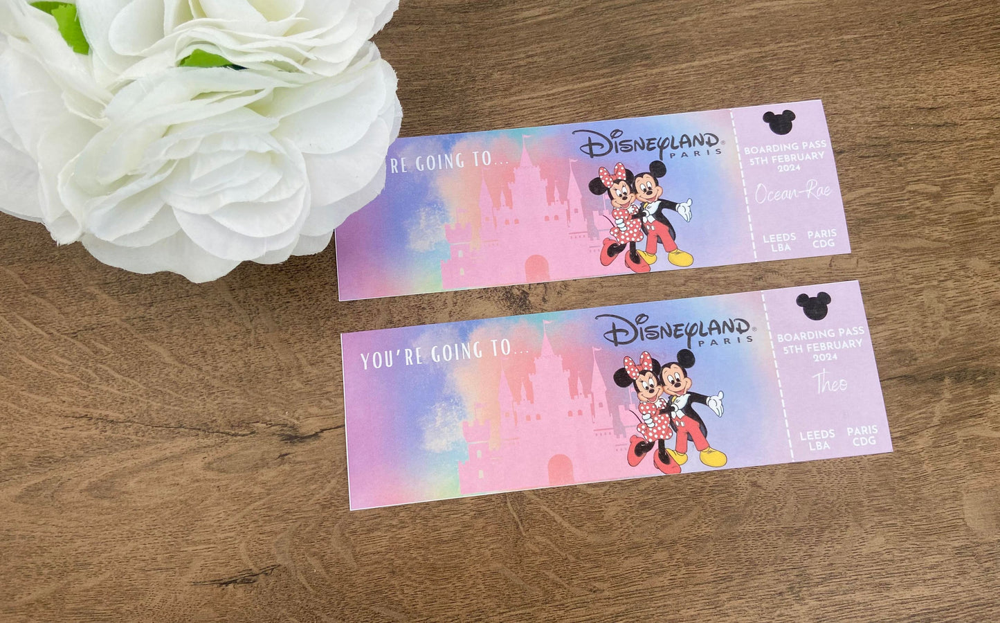 Two personalized Disney tickets with colorful graphics including Disney characters, logos, and customizable text, displayed on a wooden surface next to a white flower.