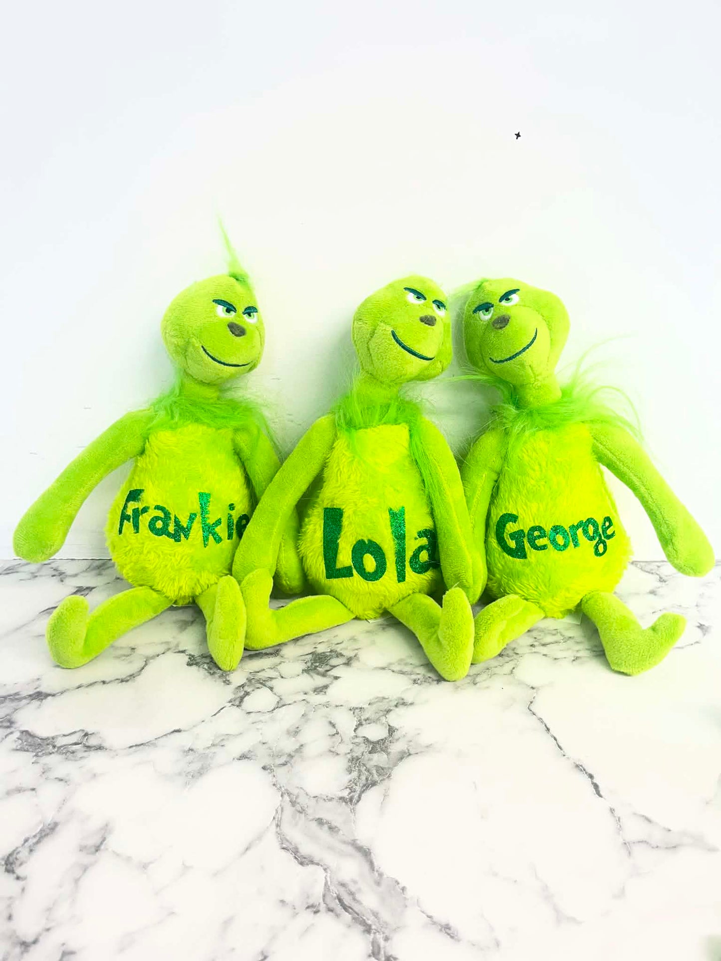 Three plush Grinch-style toys in bright green are arranged side by side on a white marble surface against a white wall. Each plush has a different name printed on the front: Frank, Lola, and George.