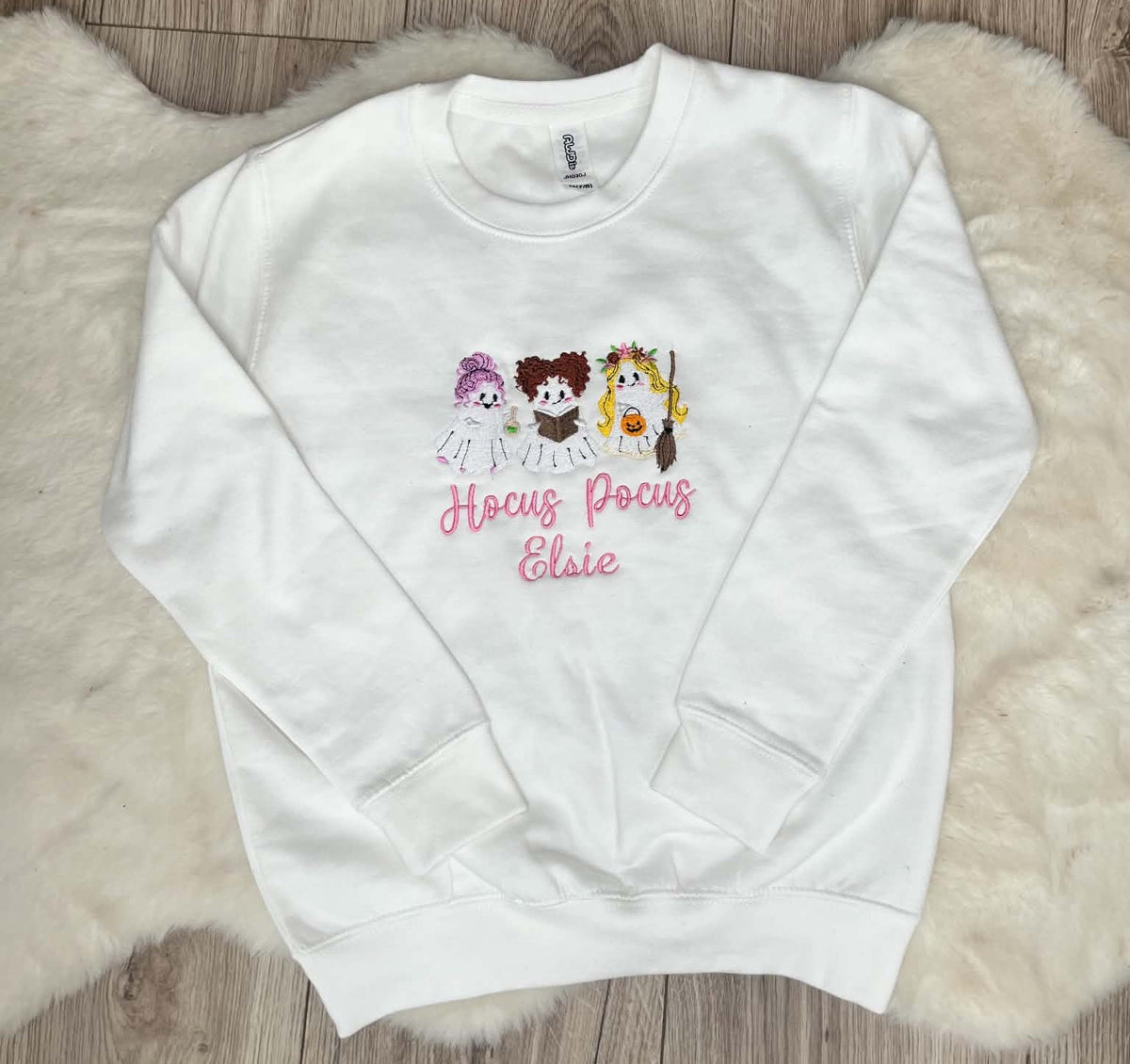 A white crewneck sweatshirt featuring a graphic of three cartoon witch characters and the text 'Hocus Pocus Ellie' in pink, laid flat on a white faux fur rug over a wood floor.