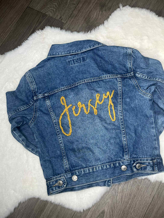 Blue denim jacket with 'Jersey' embroidered in yellow on a white fluffy surface.