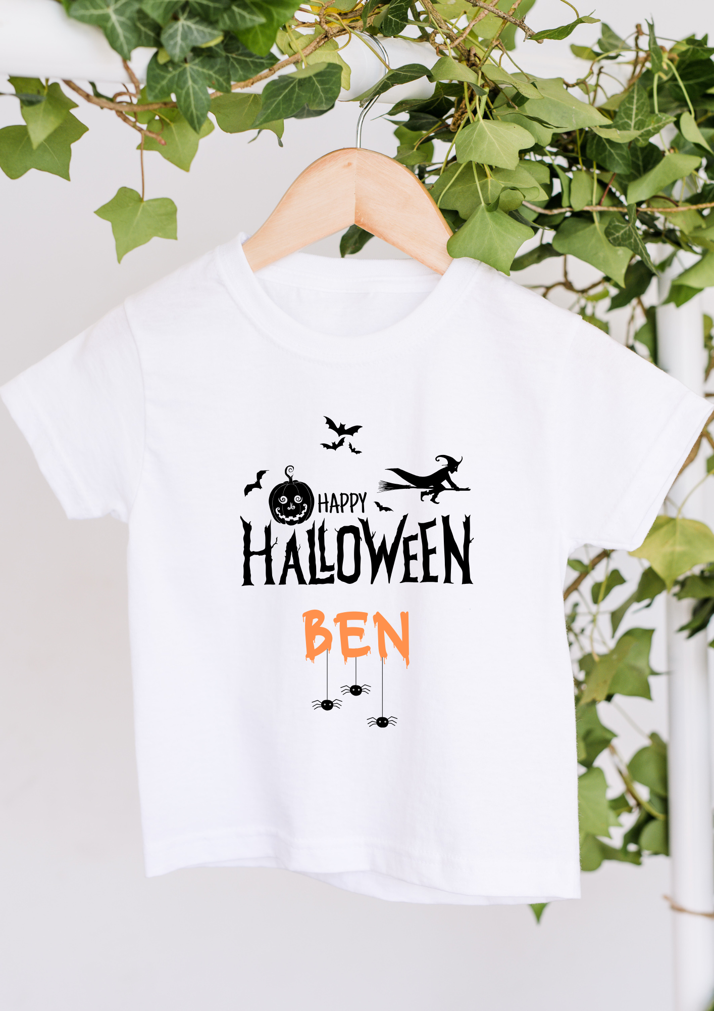 White t-shirt with 'Happy Halloween Ben' print on a hanger with greenery in the background