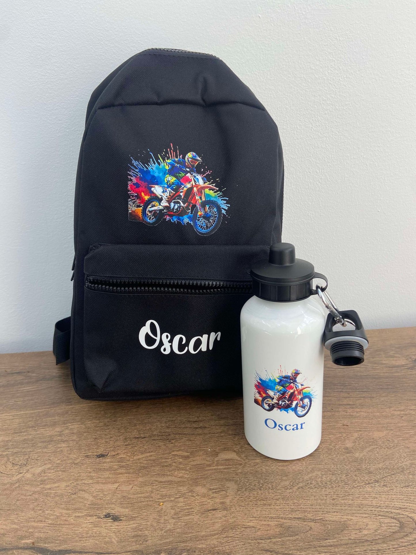 A black personalised motorbike backpack with a multicolored design on the front, a water bottle with a motorbike label, and adjustable shoulder straps, all laid out on a wooden surface.