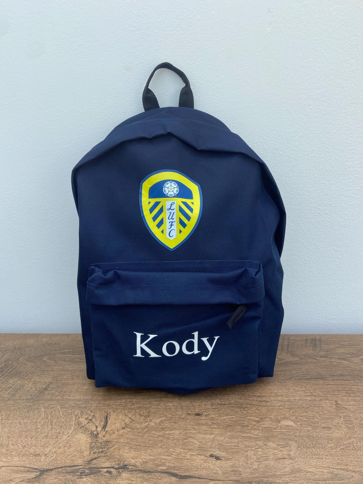 Navy blue backpack with a logo and 'Kody' text on a wooden surface and light gray background