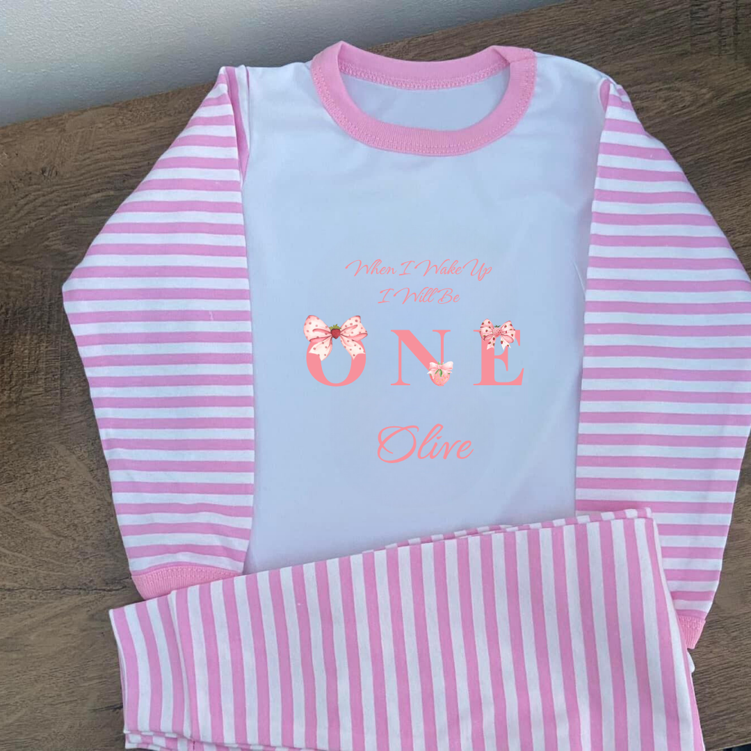 Pink and white striped children's pajama set with 'One' and name on a wooden surface