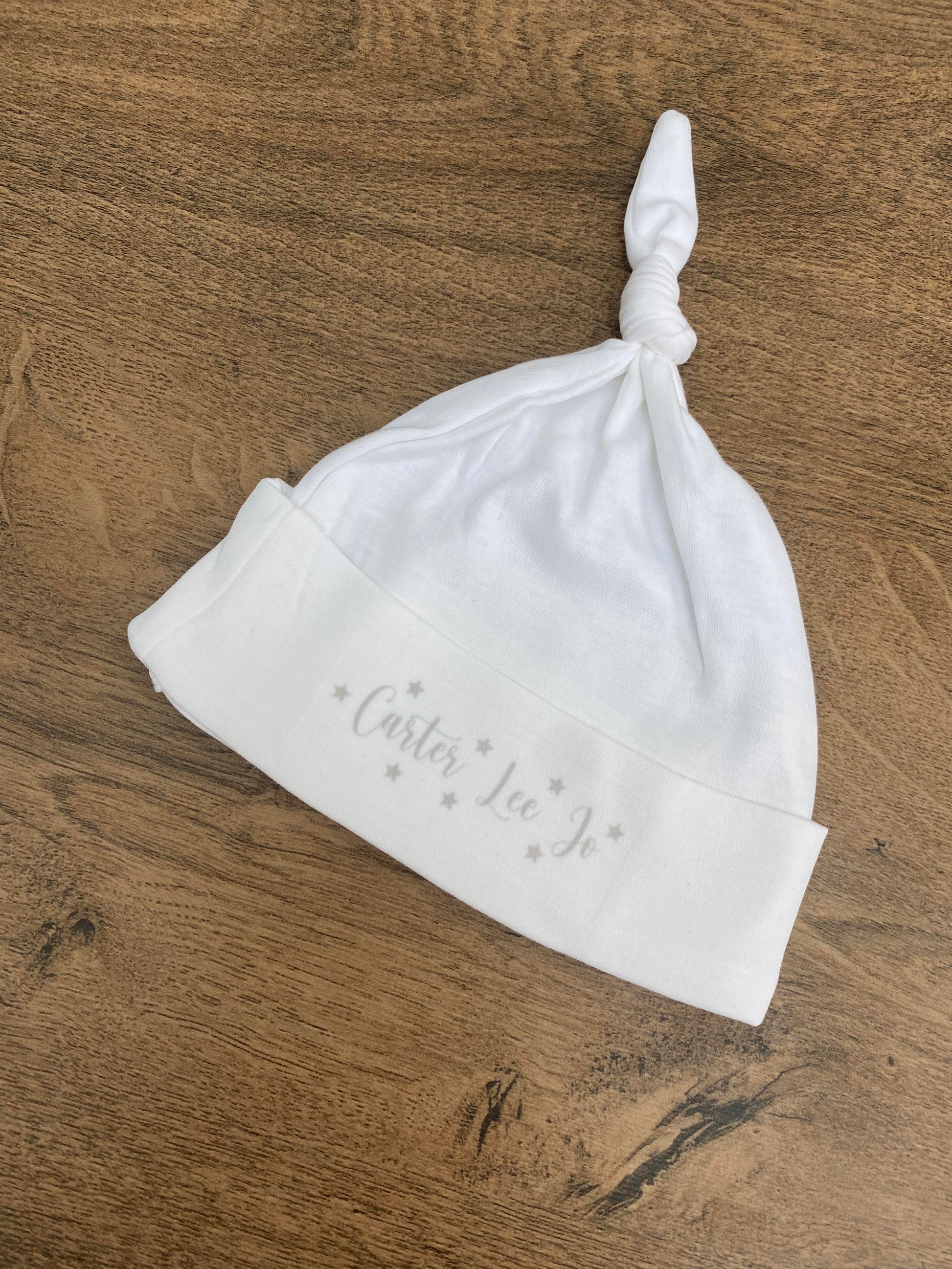 A white baby hat with a knotted top and personalised text printed on the front, placed on a wooden surface.