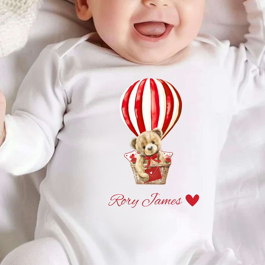 Baby wearing a white onesie with a teddy bear and hot air balloon design, featuring the name 'Rory James'.