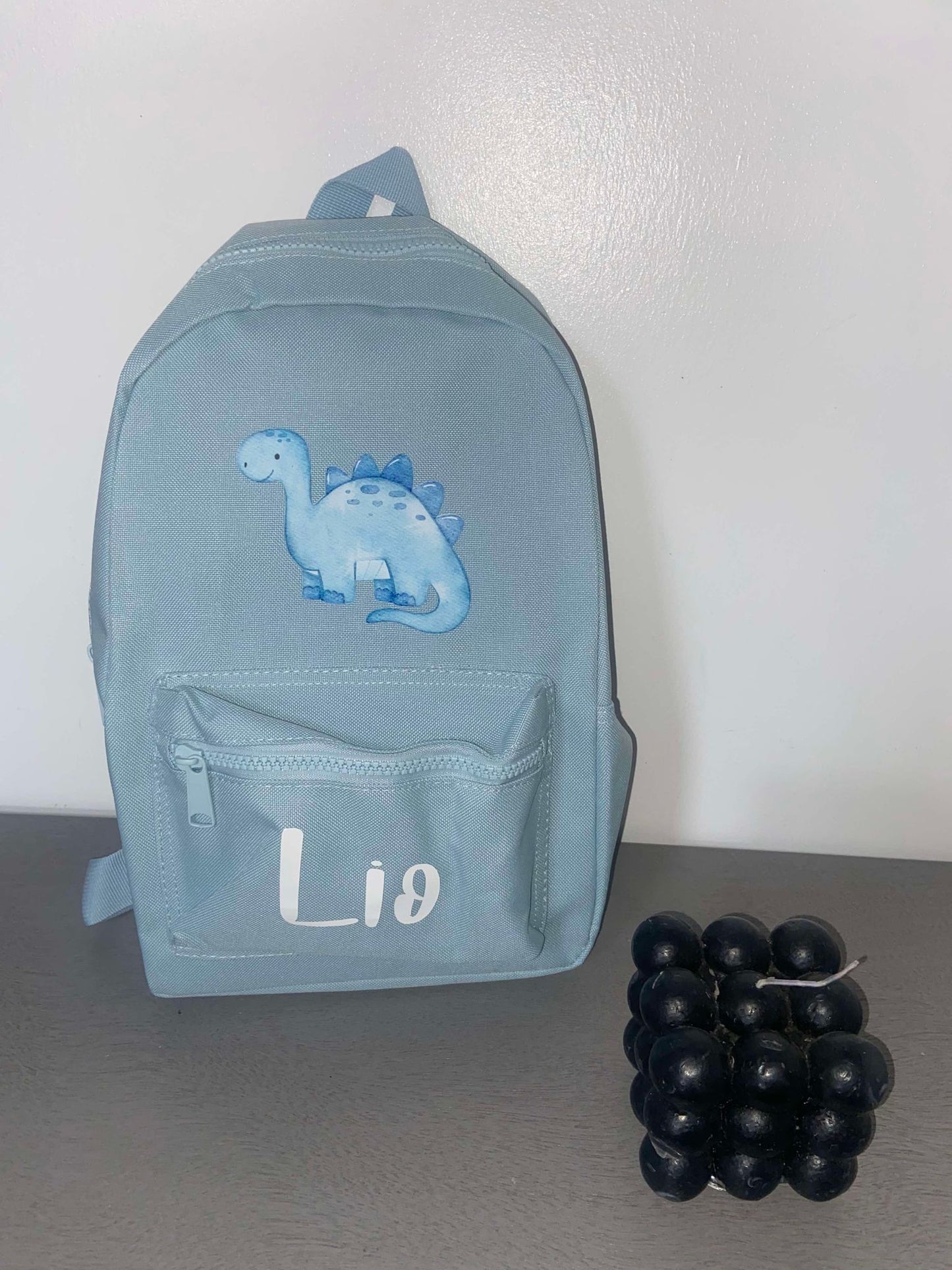 A blue personalised dinosaur-themed backpack with adjustable shoulder straps, displayed alongside a front pocket, on a surface with a few grapes.