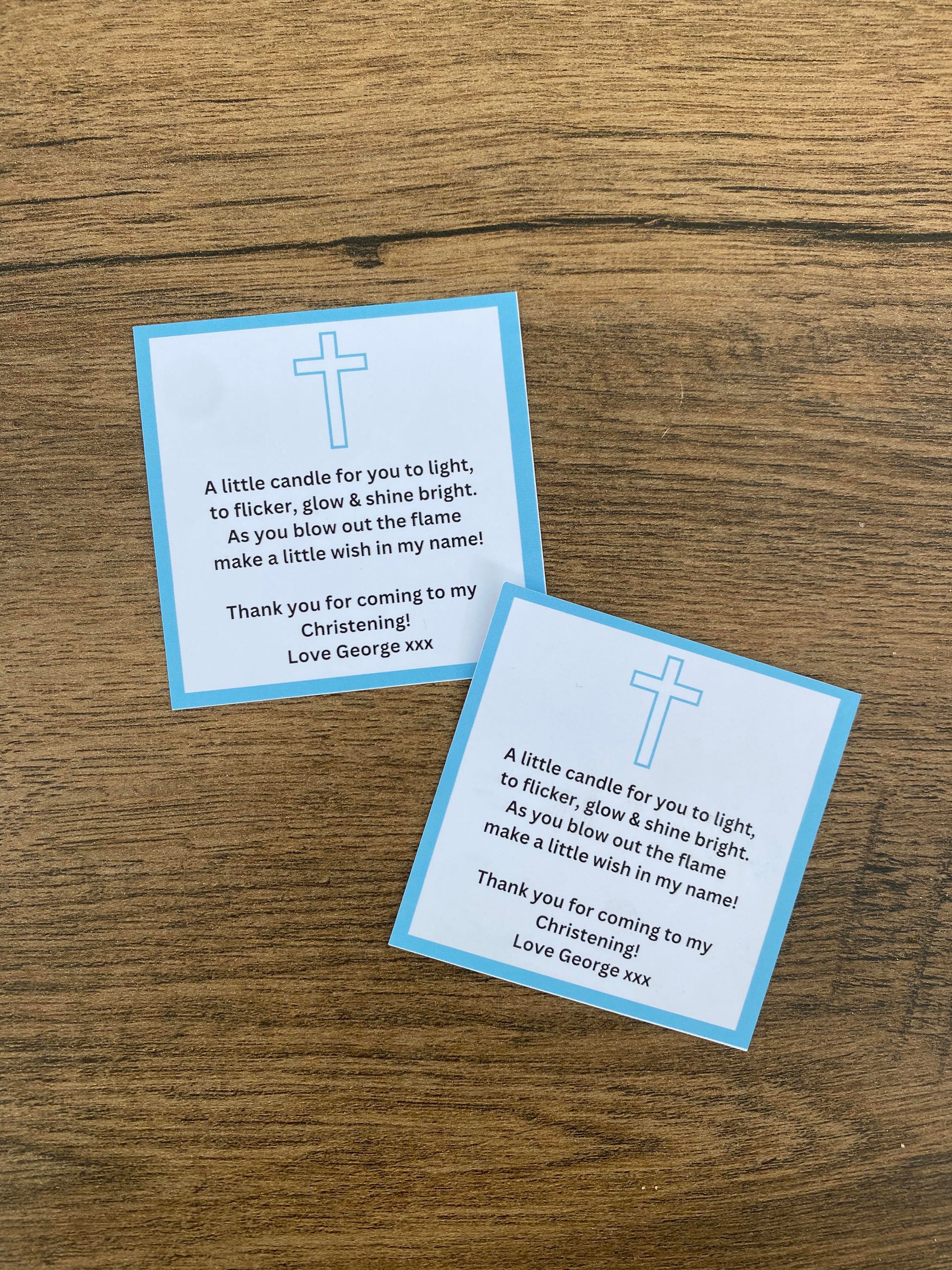 Two blue personalized Christening favour tags with white text and a cross symbol, placed on a wooden surface.