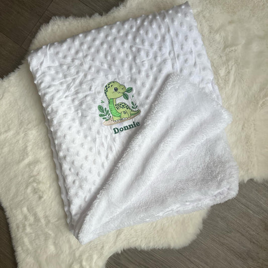 White baby blanket with a green dinosaur design and 'Donnie' text on a fluffy white surface.