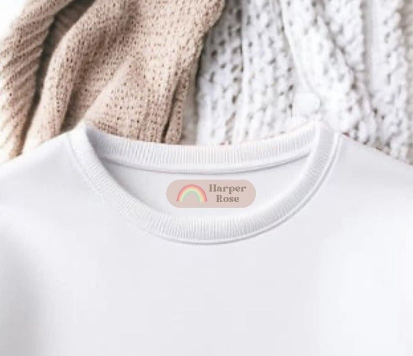 A white shirt with an iron on name label that reads 'Harper Rose' on the neckline, displayed on a cozy white background with a beige knit hat and a white blanket.