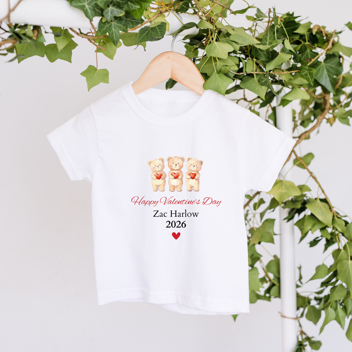 White t-shirt with Valentine's Day design featuring two bears holding hearts, text 'Happy Valentine's Day', and name 'Zac Harlow' on a hanger with green leaves in the background.