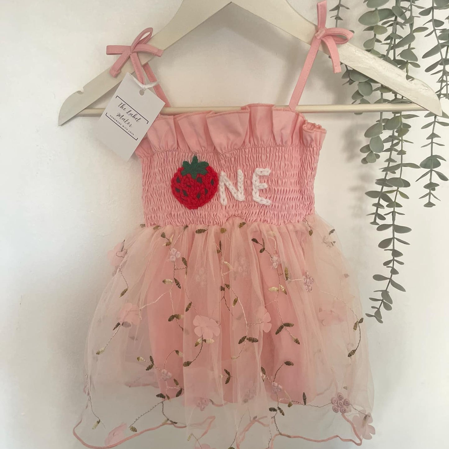 Pink baby dress with strawberry design and 'ONE' text on a hanger against a white background.
