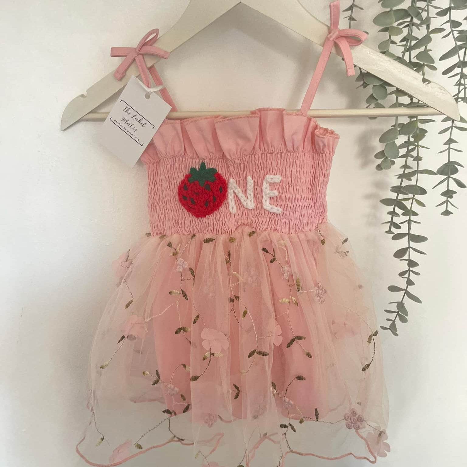 Pink baby dress with strawberry design and 'ONE' text on a hanger against a white background.