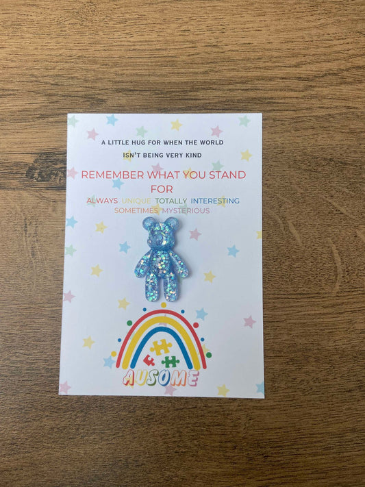 A multicolored bear-shaped pin with a rainbow and stars on a note card, likely intended as a gift or keepsake for an autistic individual.