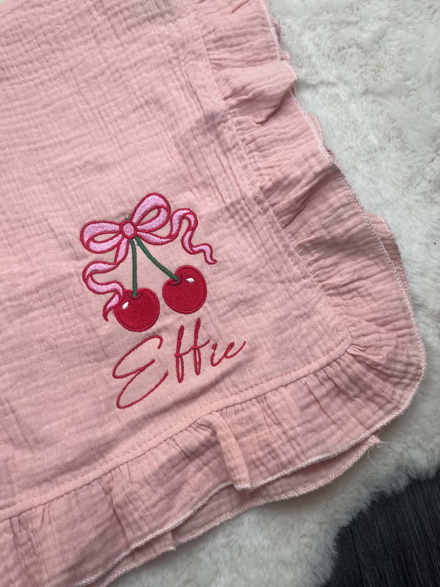 A close-up of a light pink crinkle gauze textile, likely a baby blanket or swaddle, featuring an embroidered design of two red cherries with a pink bow and the name 'Ellie' in pink script.