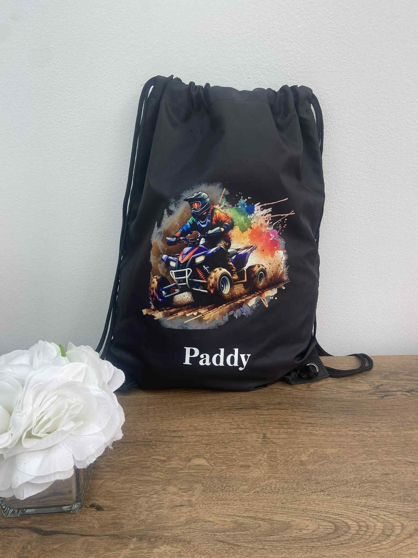 Black drawstring bag with a colorful graphic of a person on a bike and the name 'Paddy' on a wooden surface.