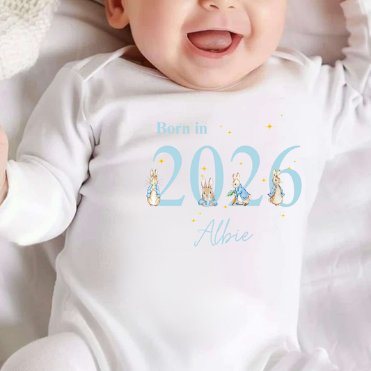 Baby wearing a white onesie with 'Born in 2026' and cartoon characters printed on it.