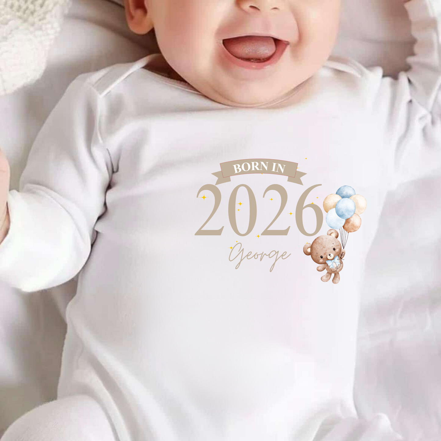 Baby wearing a white onesie with 'Born in 2026' text and teddy bear design.