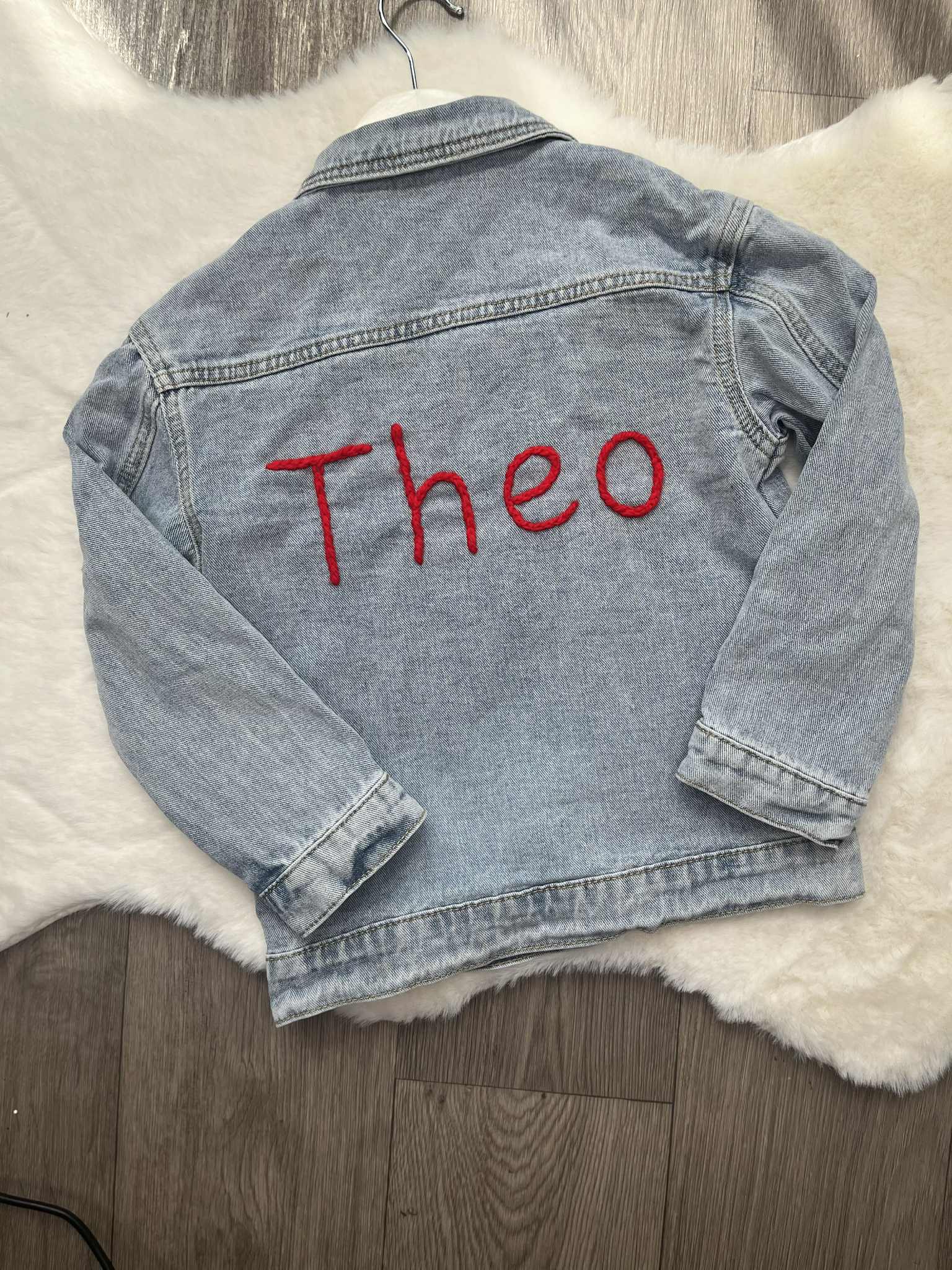 Denim jacket with 'Theo' embroidered on it, placed on a textured surface.
