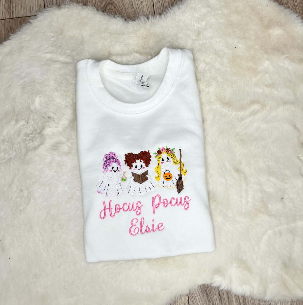 A folded white t-shirt with a graphic of three cartoon characters and the text 'Hocus Pocus Elsie' in pink, displayed on a white faux fur rug.