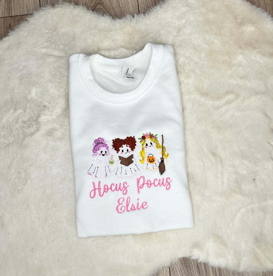 A folded white t-shirt with a graphic of three cartoon characters and the text 'Hocus Pocus Elsie' in pink, displayed on a white faux fur rug.