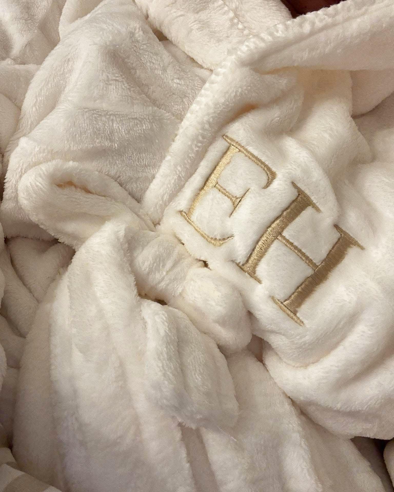 A close-up of a plush cream fleece blanket with gold embroidered initials 'EH' on a soft, textured background.