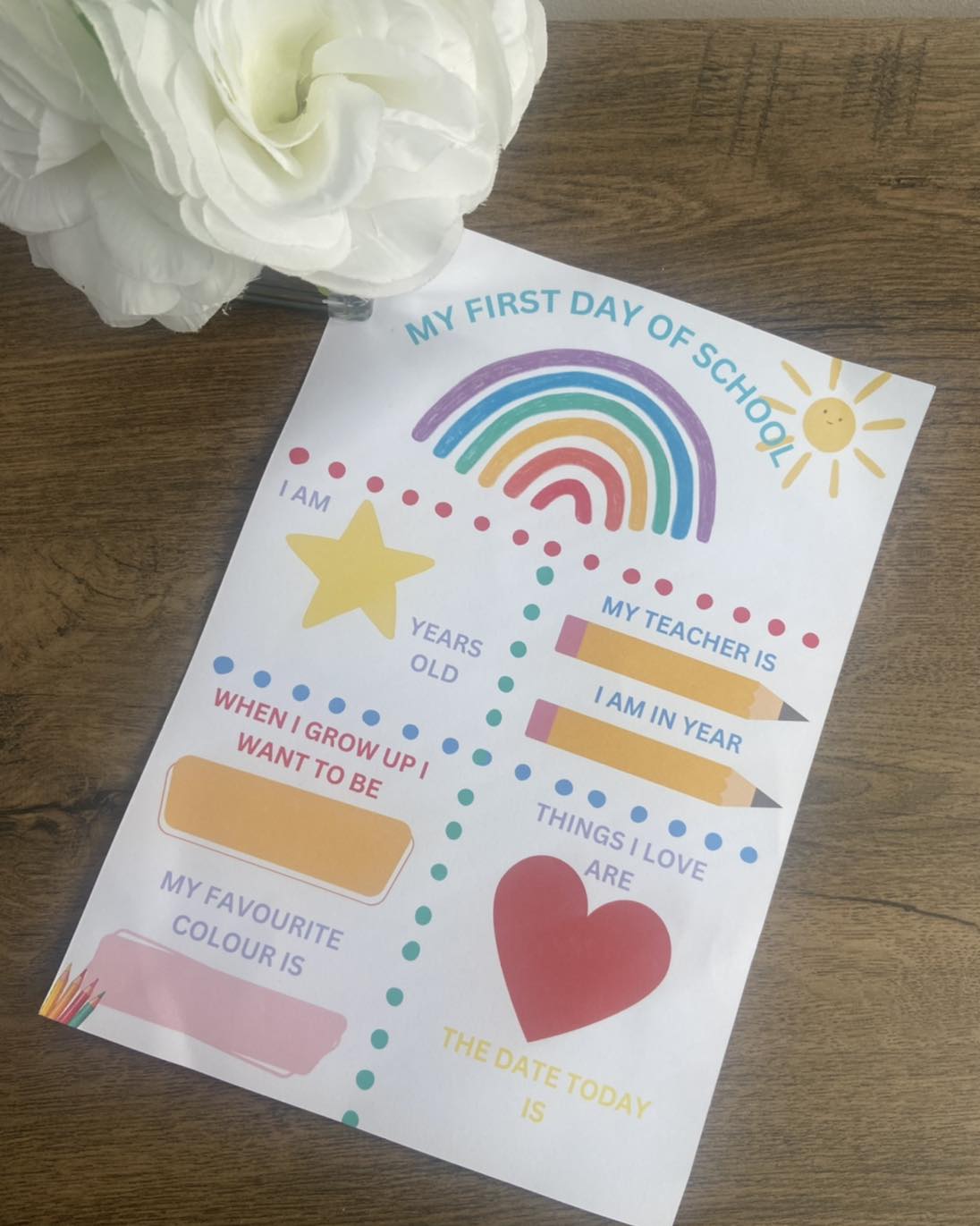 A colorful First Day of School print with spaces for writing and drawing, placed on a table with a white flower in the background.