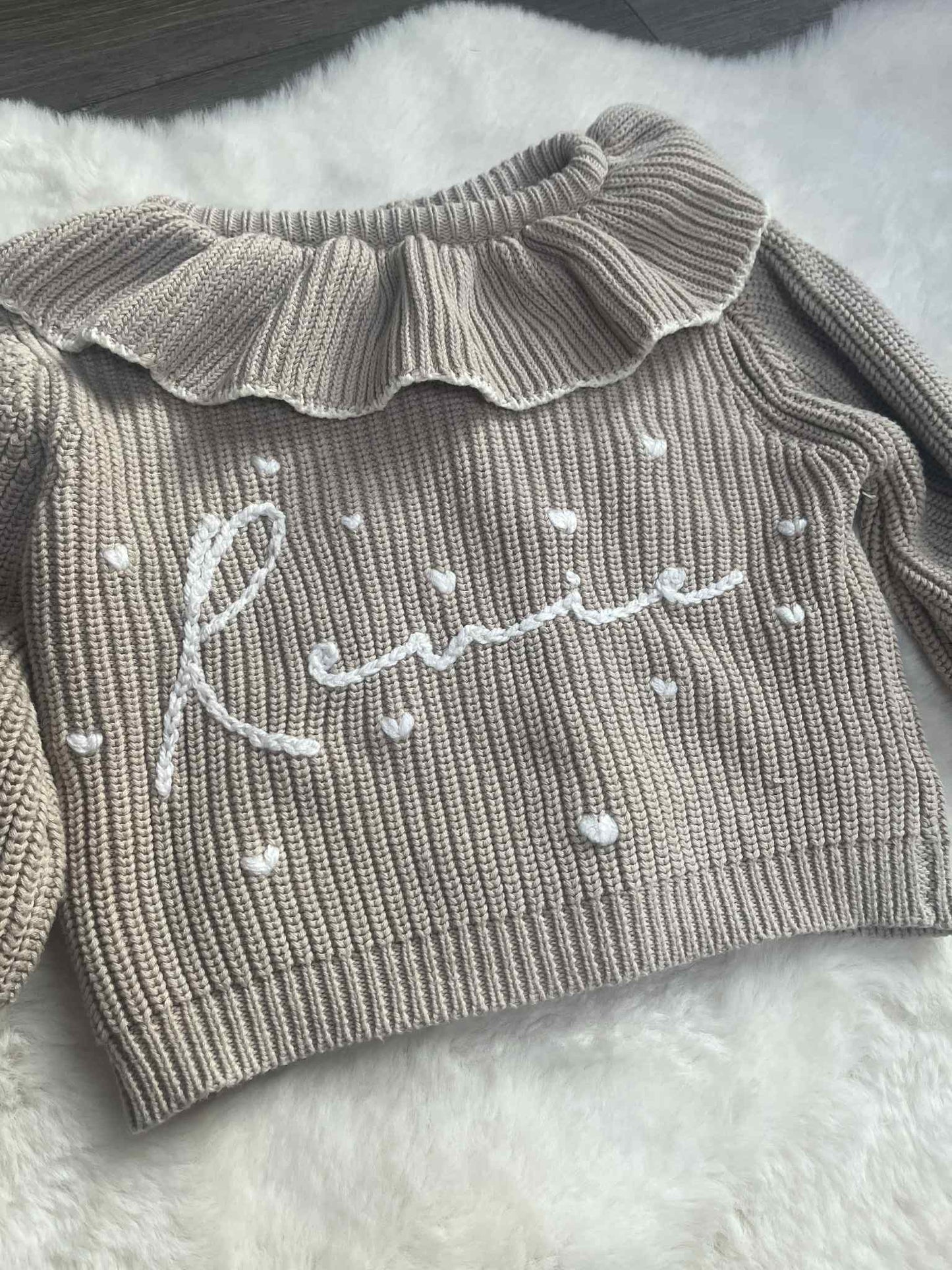 Gray ribbed sweater with ruffled collar and embroidered name on a fluffy white surface