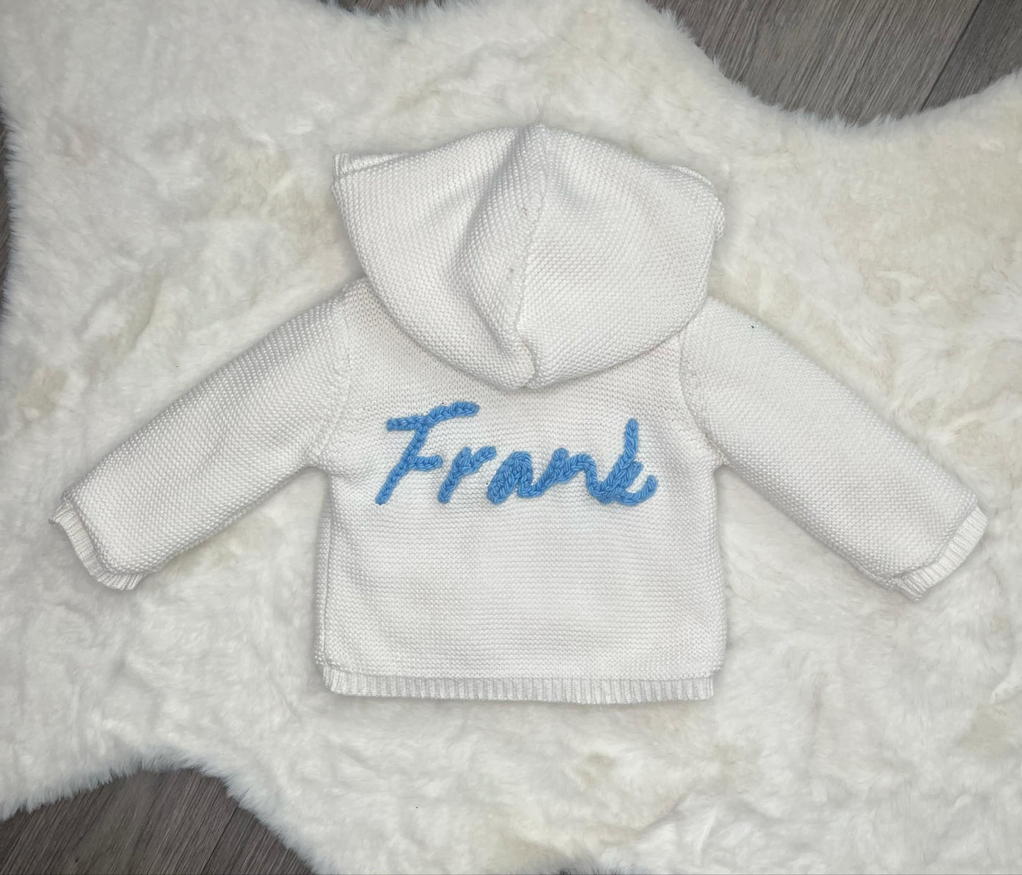 White baby kniited jacket with blue text 'Frank' on a textured white background