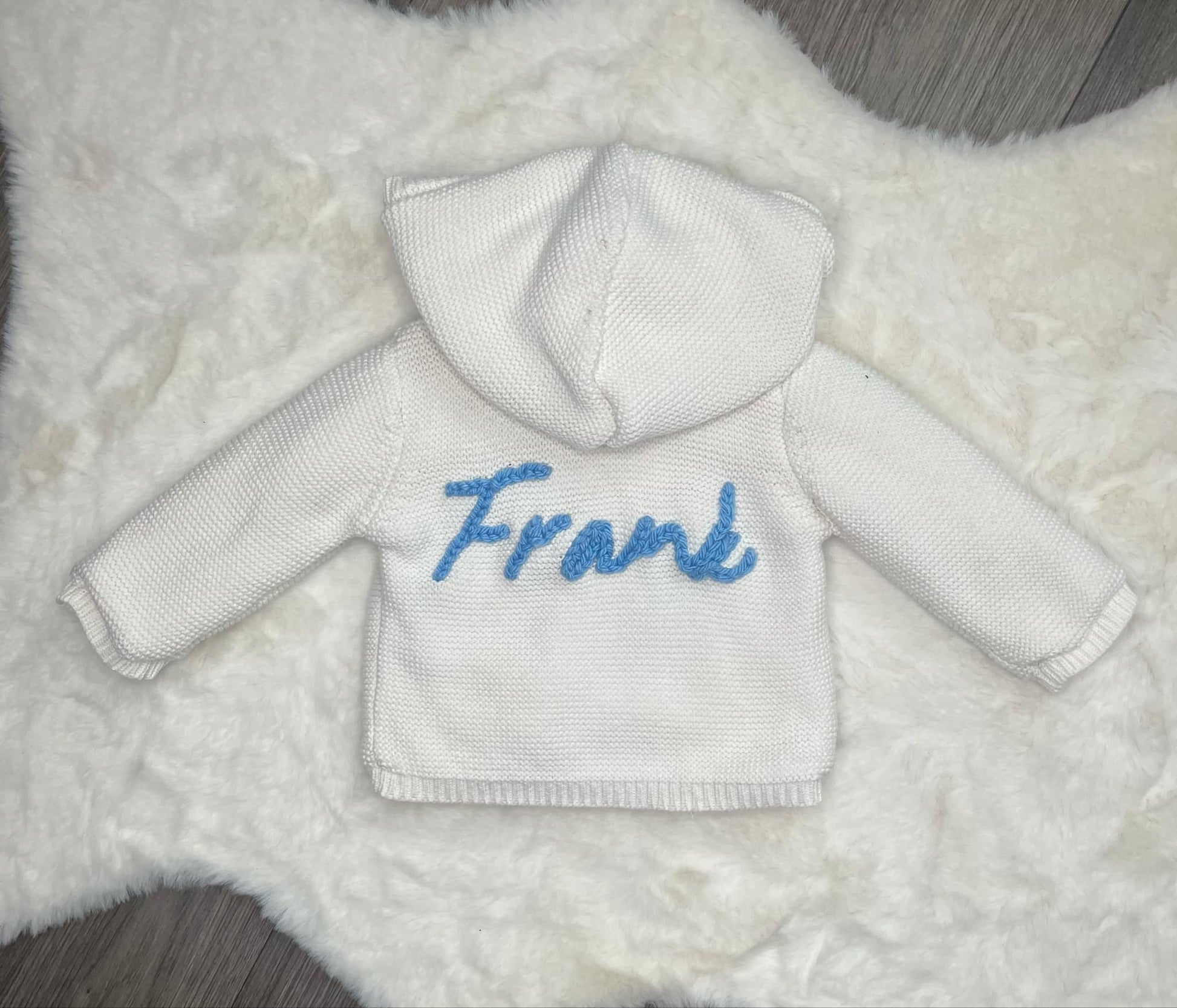 White baby kniited jacket with blue text 'Frank' on a textured white background