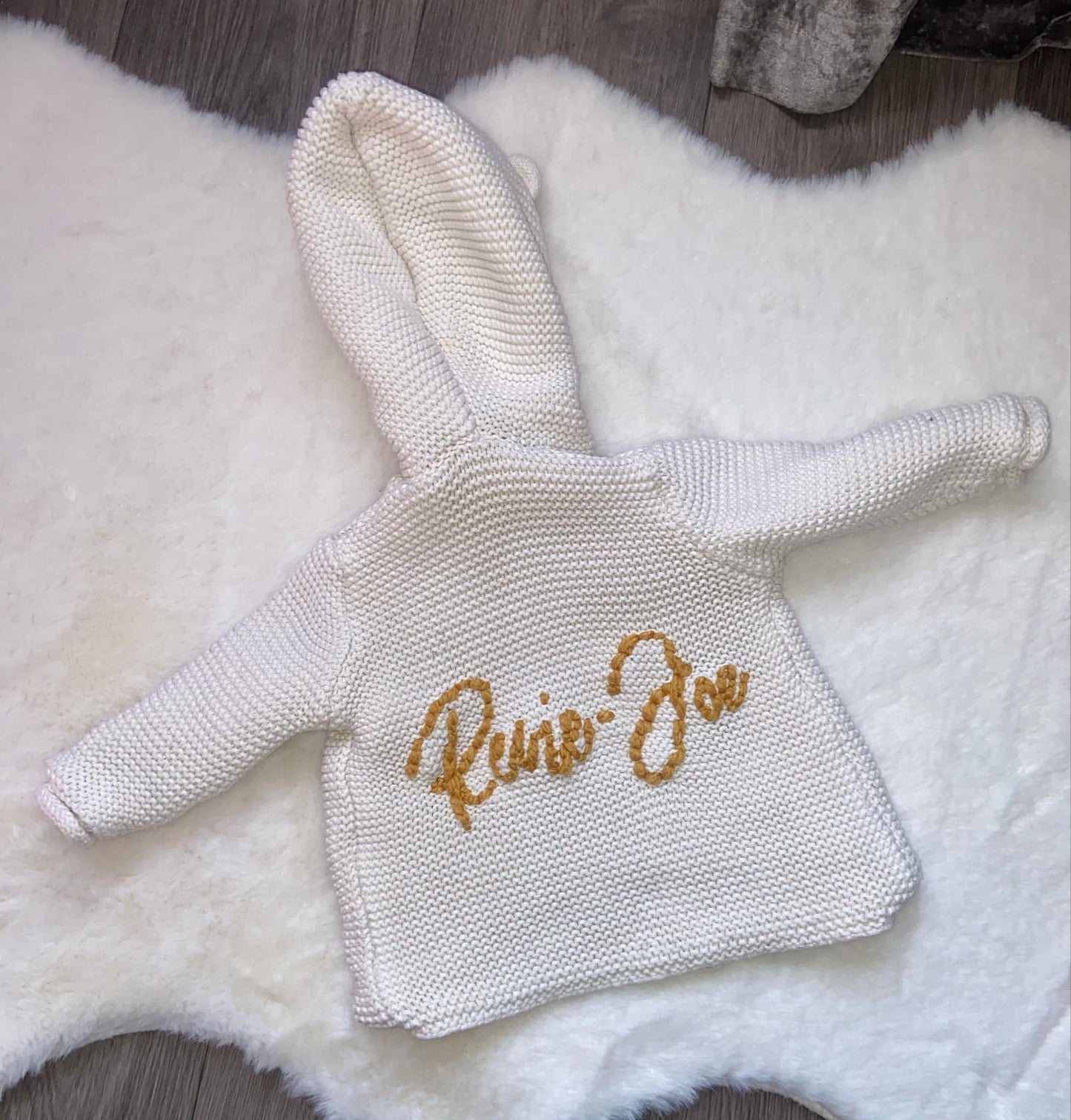 Knitted baby outfit with 'Raine Zoe' embroidery on a fluffy white surface.