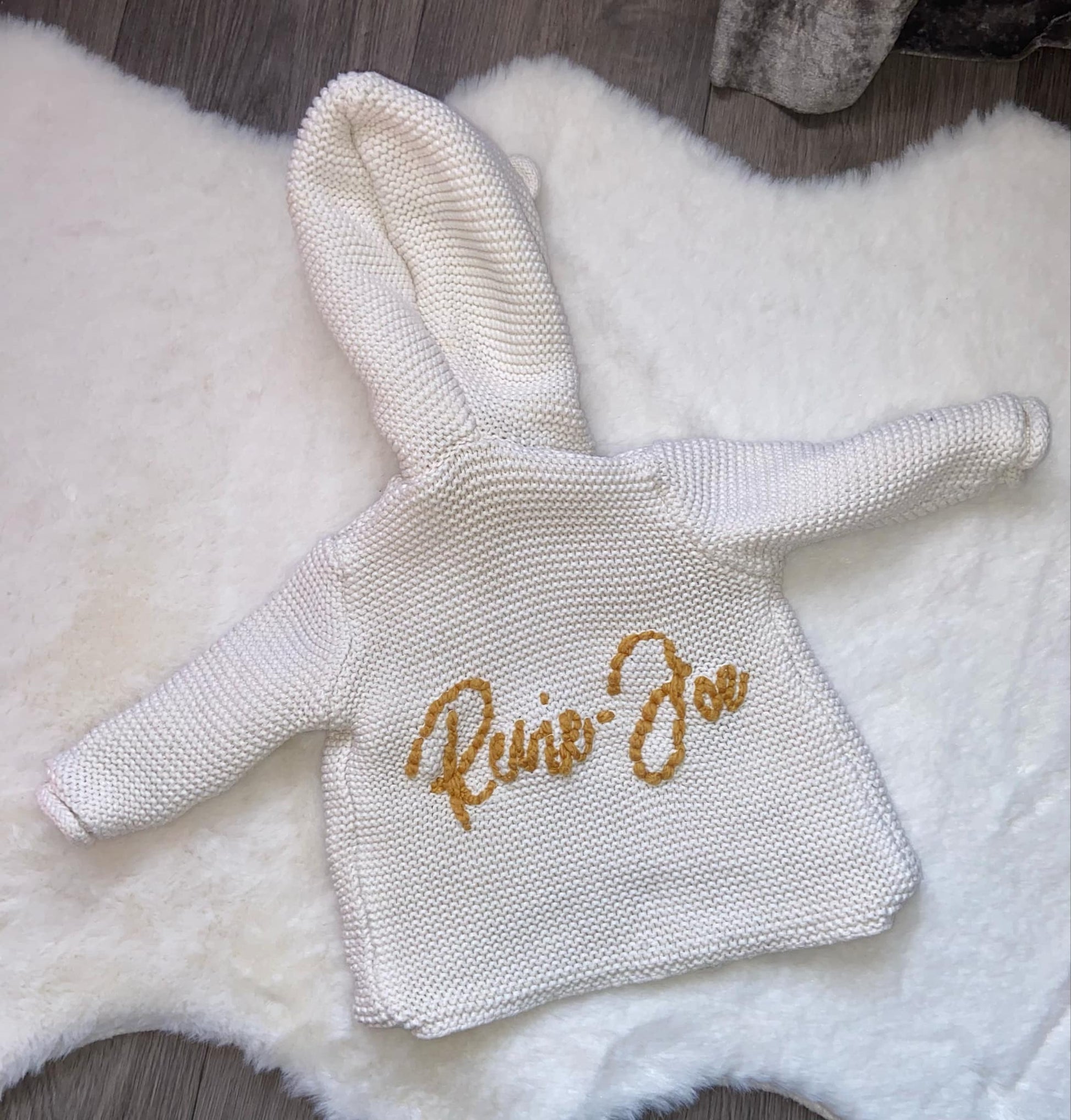 Knitted baby outfit with 'Raine Zoe' embroidery on a fluffy white surface.