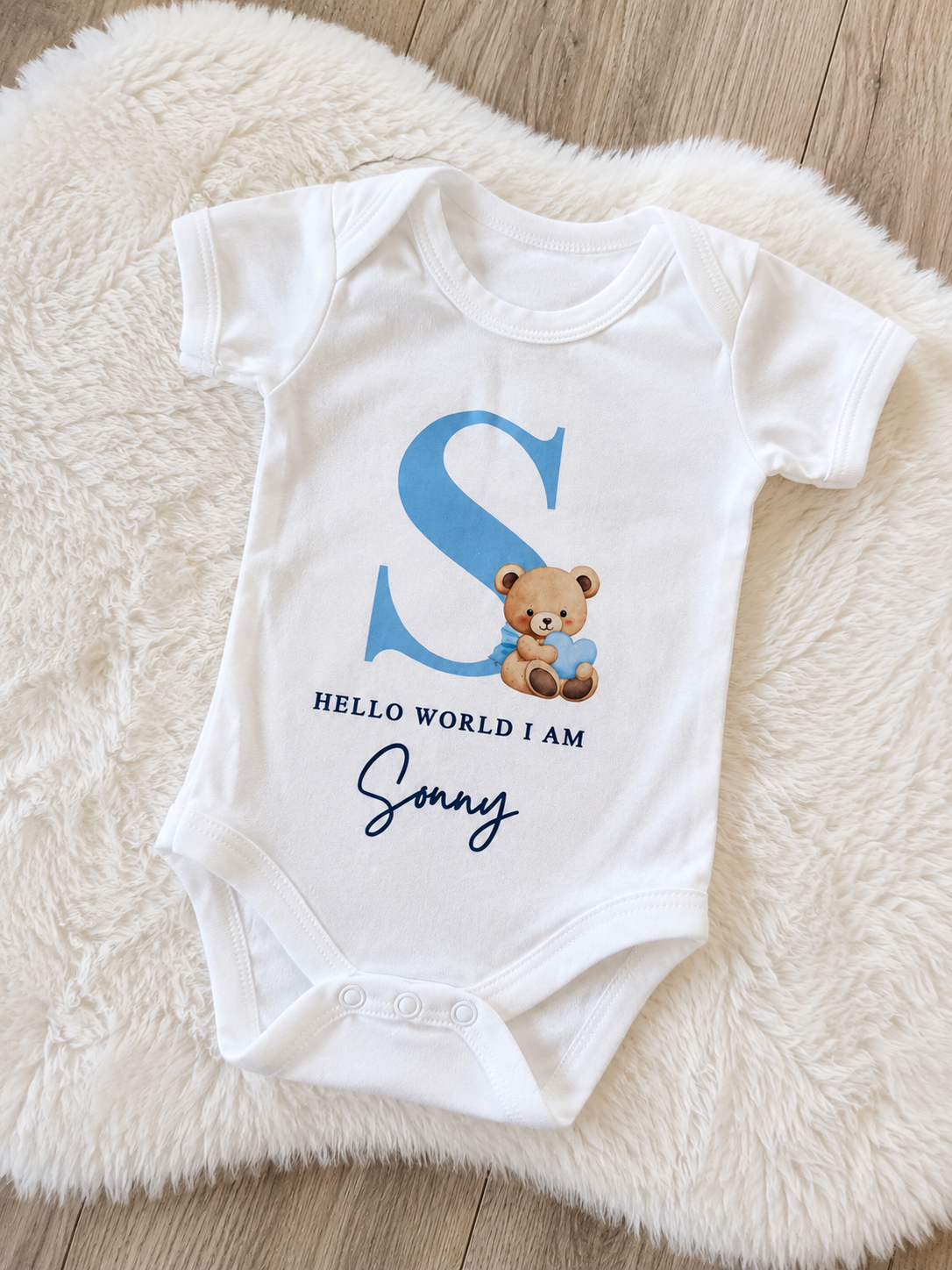 White baby onesie with blue letter 'S', teddy bear, and text on a fluffy white surface.
