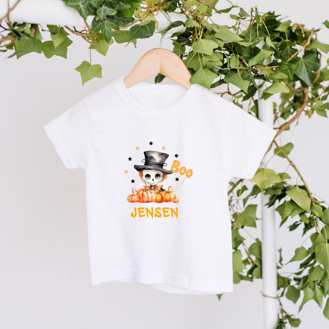 White t-shirt with Halloween design featuring a skull, pumpkins, and text on a hanger with green leaves in the background.