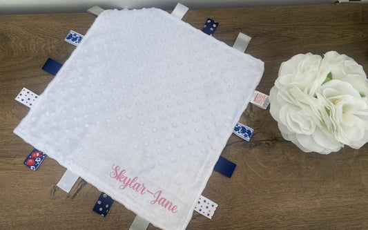 A personalised white baby comforter with blue and red patches, and a custom name printed in pink.