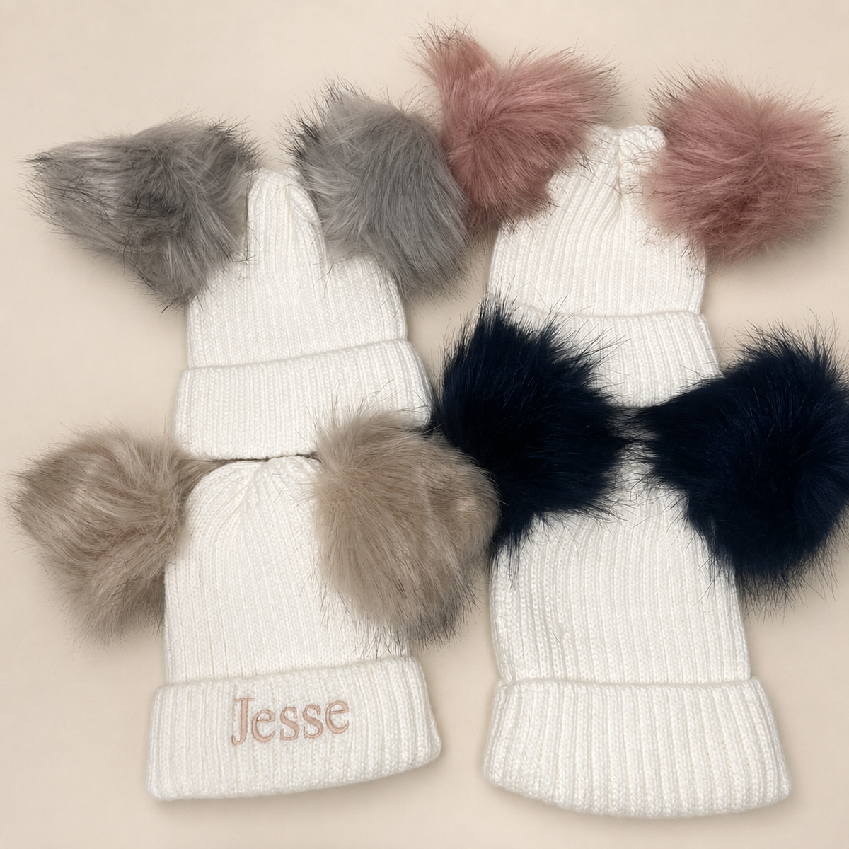 Three white ribbed knit baby beanies with faux fur pom-poms, arranged on a light beige background. One beanie has a pink embroidered name 'Jesse' on the cuff, and the pom-poms are gray, blush pink, and dark navy.