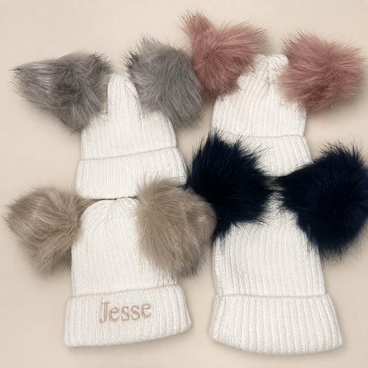Three white ribbed knit baby beanies with faux fur pom-poms, arranged on a light beige background. One beanie has a pink embroidered name 'Jesse' on the cuff, and the pom-poms are gray, blush pink, and dark navy.