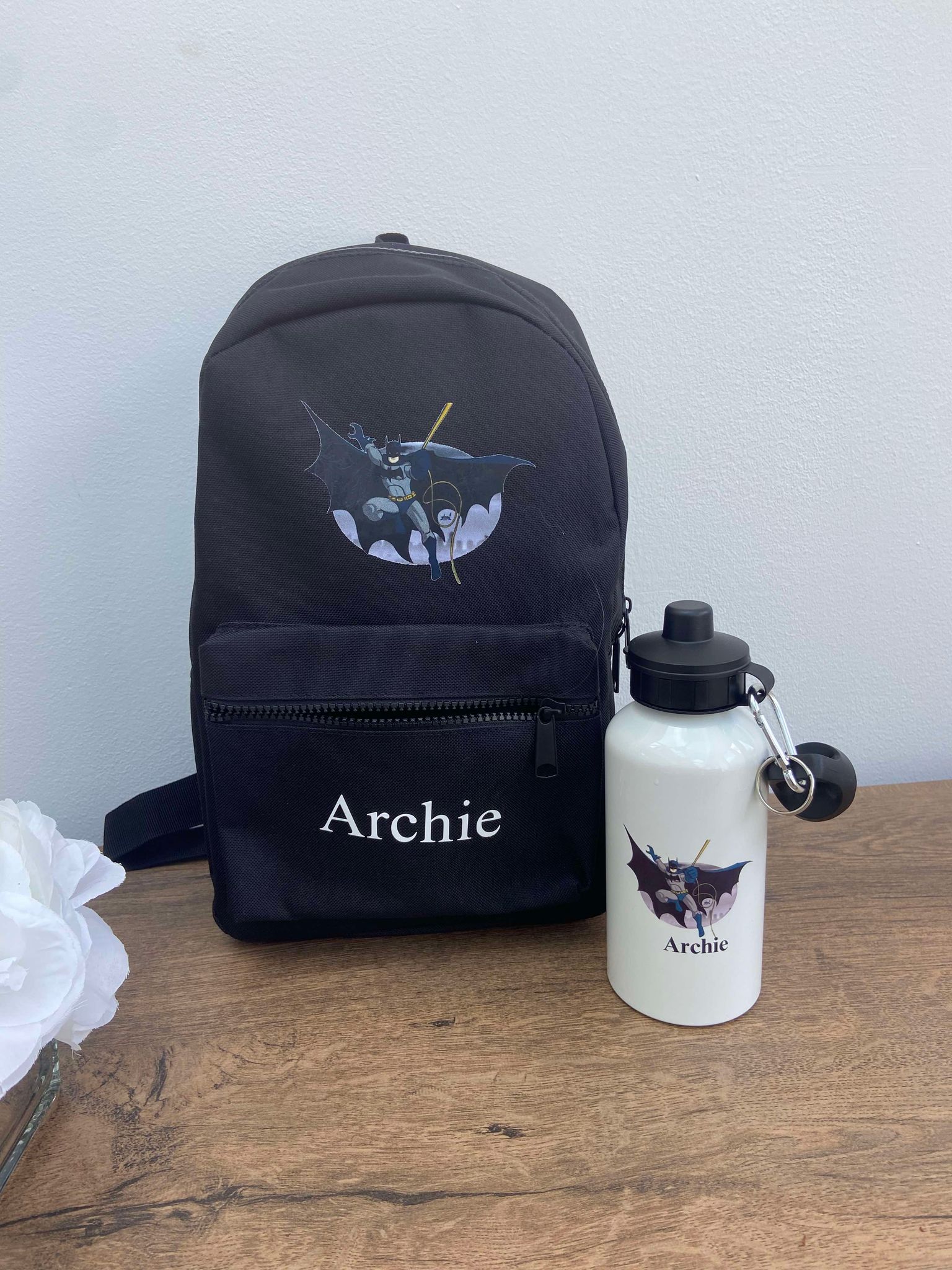 A black personalised backpack for children with a Bat Man design, accompanied by a white water bottle with a name 'Archie' printed on it.