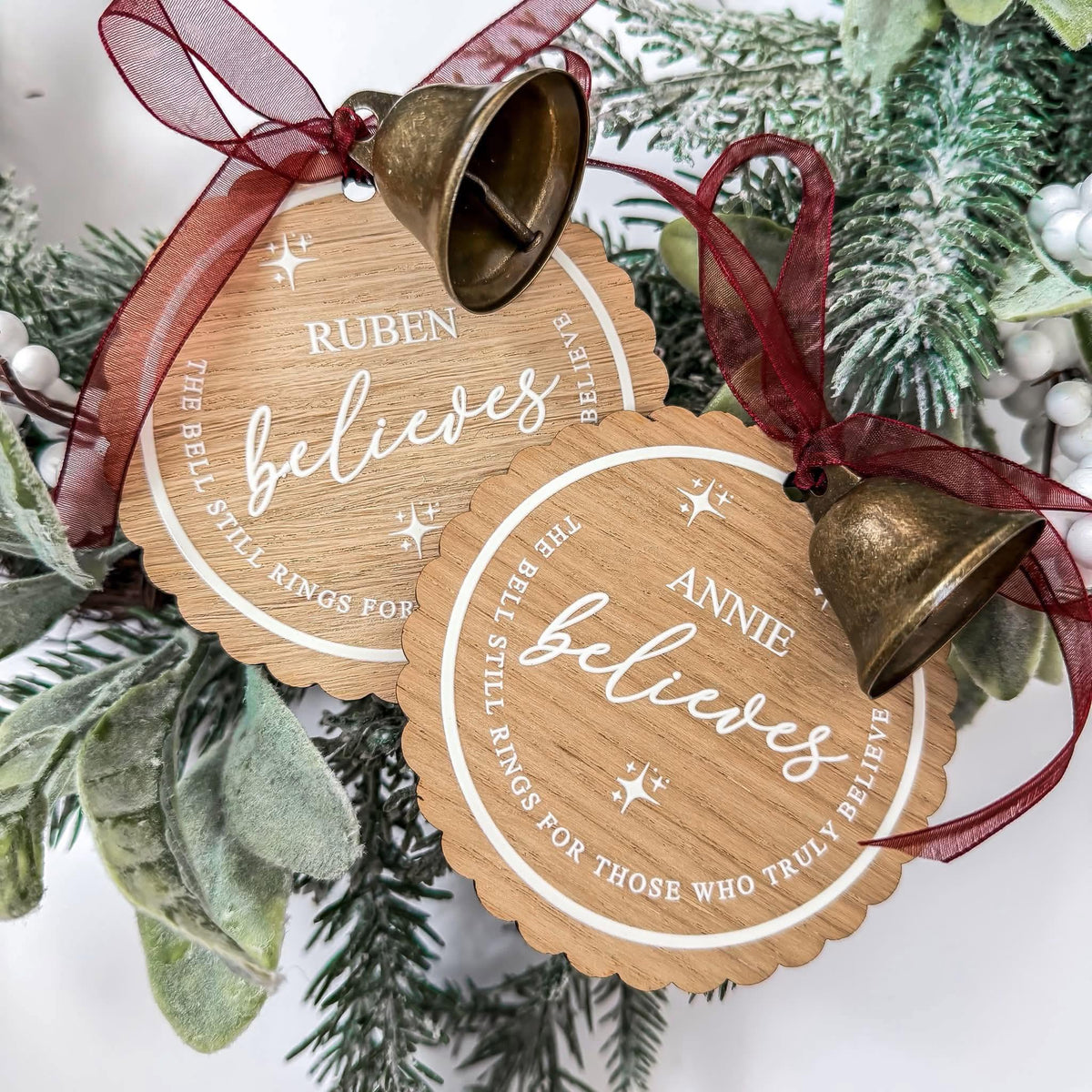 Two personalized wooden Christmas ornaments with hanging bronze bells and red ribbon, displayed among green pine branches and white berries. The ornaments are engraved with names, 'believes,' and a quote about the bell ringing for those who truly believe.