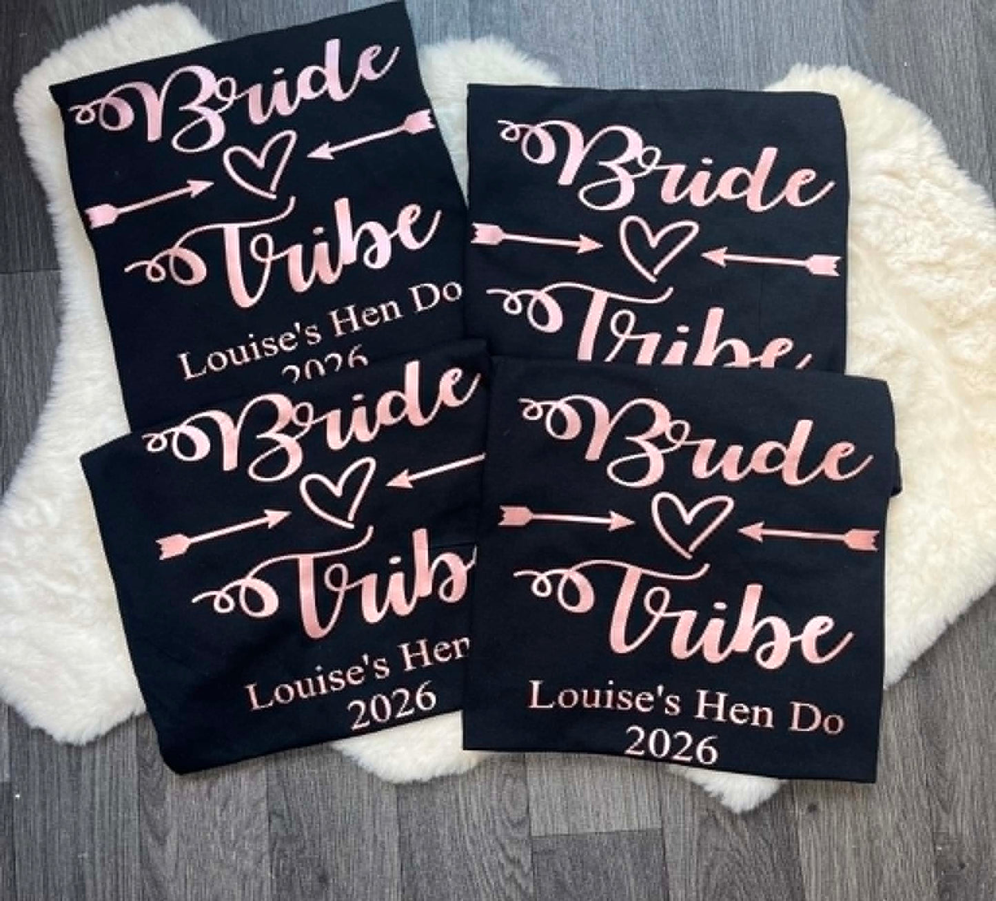 Four black fabric prints laid flat on a white faux-fur rug over a gray wood floor. Each print features white script reading 'Bride Tribe' with a heart and arrows, plus personalized text 'Louise's Hen Do 2026' in white and light pink.