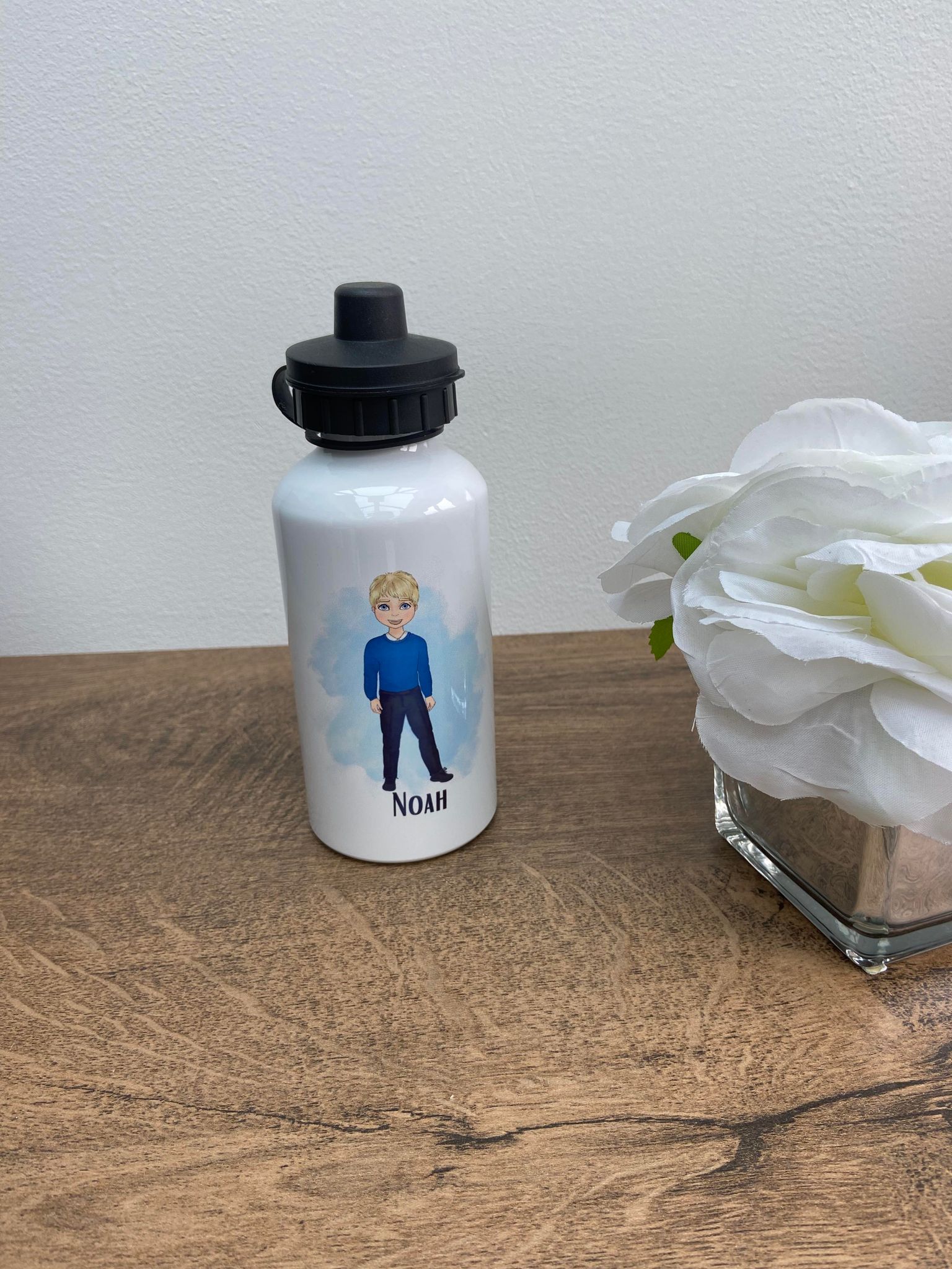 A white water bottle with a custom character design, featuring a child with blonde hair in a blue uniform, placed on a wooden table next to a white flower decoration.