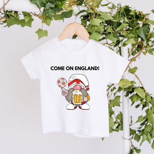 White t-shirt with a cartoon character and 'COME ON ENGLAND!' text, hanging against a white background with greenery.