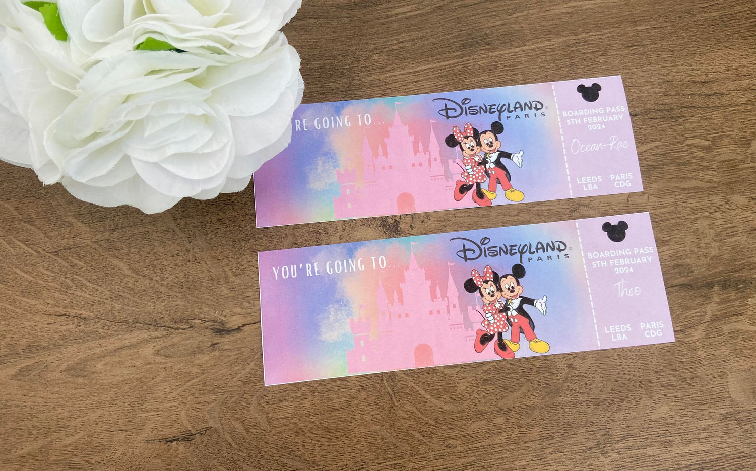 Two personalized Disney tickets with colorful graphics including Disney characters, logos, and customizable text, displayed on a wooden surface next to a white flower.