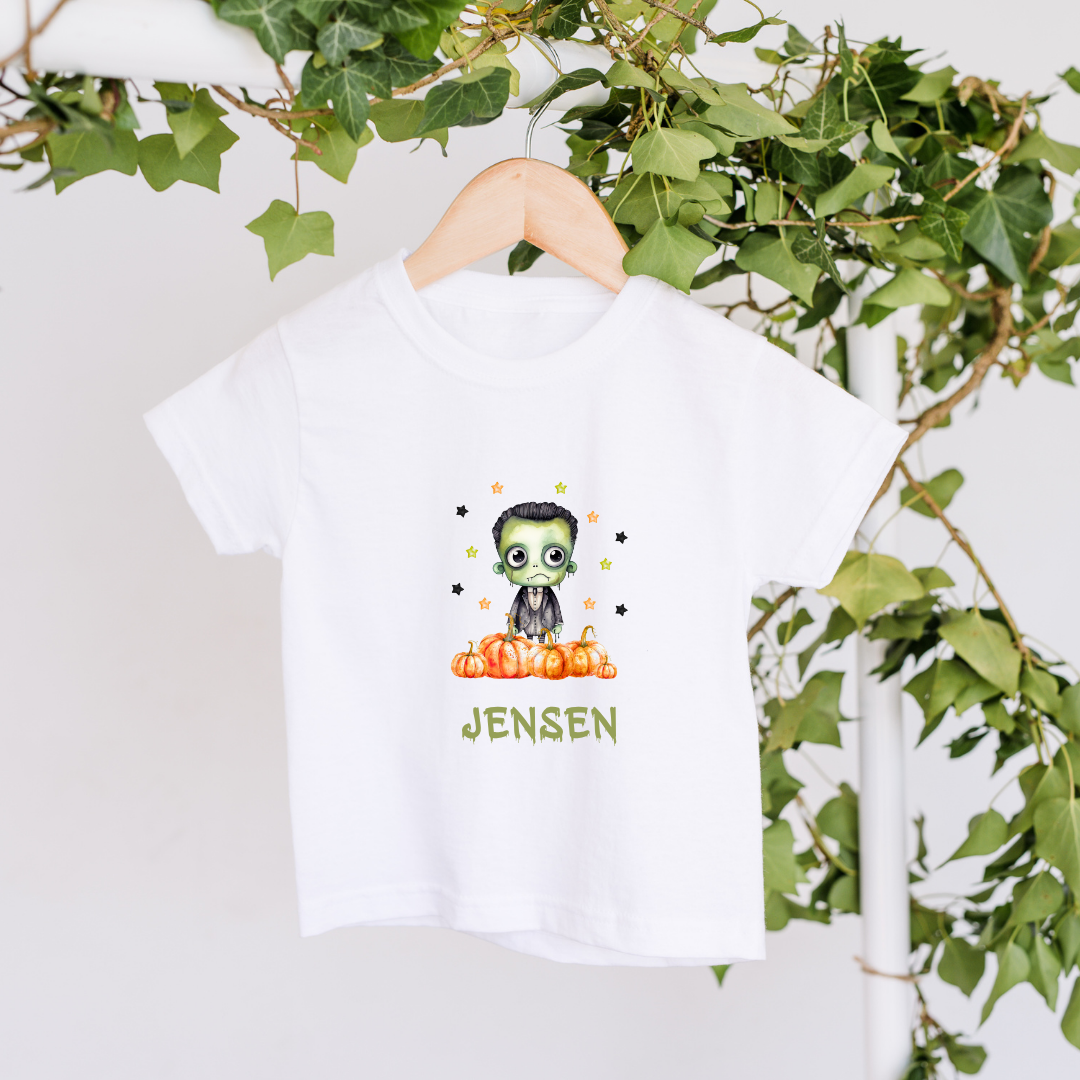 White t-shirt with a graphic design of a character in a suit and pumpkin, with 'JENSEN' printed below, on a hanger with green leaves in the background.