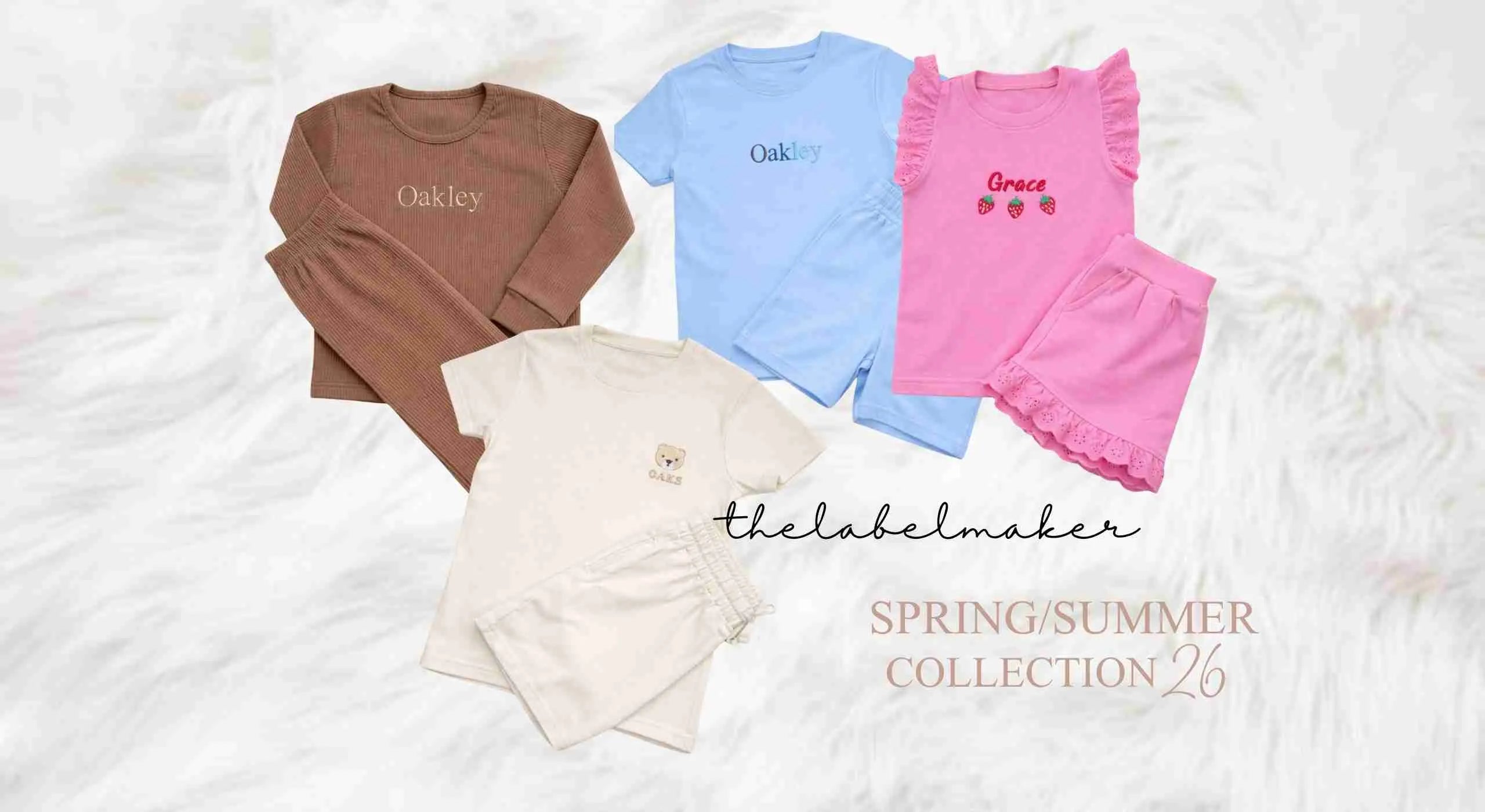 Four children's two-piece outfits arranged on a white textured fabric background. The set includes a brown long-sleeve top with matching shorts, a light blue short-sleeve top with matching shorts, a pink ruffled sleeveless top with matching ruffled shorts, and a cream short-sleeve top with matching shorts.