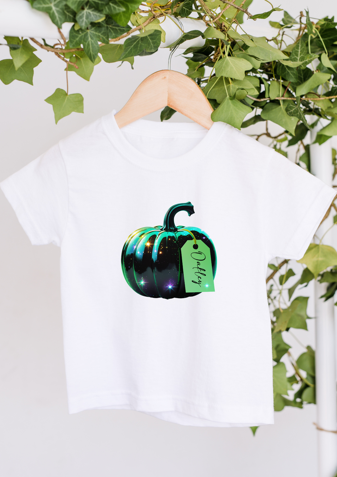 White t-shirt with a black and green pumpkin design on a hanger against a white background with greenery.
